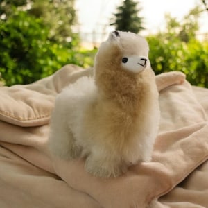 Op de afbeelding: Een pluche alpaca speelgoed van lichtbruin, zit op een crèmekleurige deken. De alpaca heeft een wit gezicht en een zachte, knuffelige uitstraling.