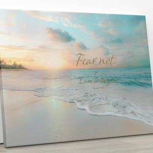 Peut inclure: Impression sur toile représentant une scène de plage sereine avec un coucher de soleil doré, une eau turquoise et du sable blanc. Le texte "Fear not, for I am with you" est écrit en script doré, ajoutant une touche de motivation.