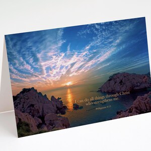 May include: Greeting card featuring a scenic sunset over the ocean with rocky cliffs. The sky is a mix of blue, orange, and pink hues. Text on the card reads: "I can do all things through Christ who strengthens me." Philippians 4:13.