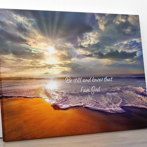 Può includere: Una stampa su tela con una scena di spiaggia al tramonto. Il cielo è un mix di rosa, arancione e blu con nuvole. Le onde si infrangono sulla riva e la sabbia è di colore dorato. Il testo "Be still and know that I am God" è stampato sulla tela.