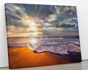 May include: A canvas print with a beach scene at sunset. The sky is a mix of pink, orange, and blue with clouds. The waves are crashing on the shore, and the sand is a golden color. The text "Be still and know that I am God" is printed on the canvas.