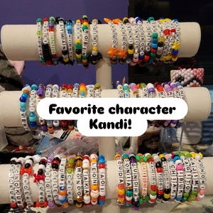 May include: A collection of colorful beaded bracelets with different names and words spelled out on them. The bracelets are displayed on a white stand. The text "Favorite character Kandi!" is written on a white sticker.