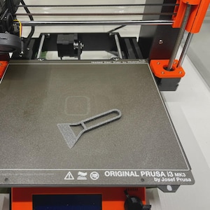 Może przedstawiać: Drukarka 3D z pomarańczowymi i czarnymi elementami. Szara skrobaczka spoczywa na podgrzewanym stole. Drukarka jest oznaczona jako "ORIGINAL PRUSA i3 MK3 by Josef Prusa" i ma etykietę "Print area".