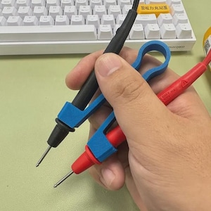 May include: A black and red multimeter test lead set with blue handles. The test leads are held in a hand, with the tips pointing towards the viewer. The background includes a white keyboard and a light green surface.