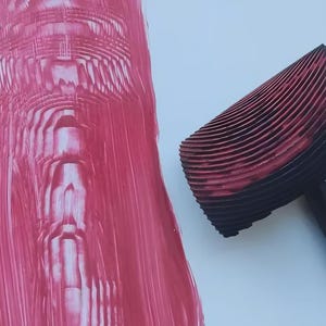 May include: A black and red patterned paint roller with a wooden handle, next to a pink painted surface. The roller has a curved, ridged design, creating a textured effect on the pink paint. The image shows a close-up of the tool and its application.