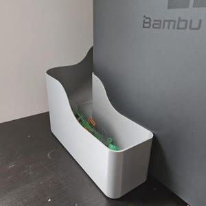 May include: A light grey storage container with a curved top. The container is empty, with a few green and orange items inside. The words "Bambu La" are printed on the grey wall behind the container.