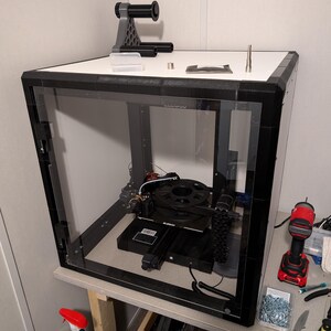 May include: A black 3D printer enclosed in a clear acrylic case with a white top panel. On the top panel sits a grey and black filament spool holder, a small clear container, a metal nozzle, and a small black packet. The printer has a black frame.