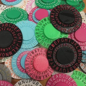 May include: A collection of colorful, circular alphabet stamps in various colors, including black, pink, green, and blue. Each stamp features a central disc and a surrounding ring of letters.