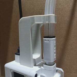 Puede incluir: Un soporte blanco impreso en 3D sujeta un tubo transparente y un componente rectangular blanco. El soporte está fijado a un dispositivo blanco con un cable de alimentación negro. La imagen se toma sobre un fondo neutro.