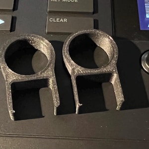 May include: Two identical black 3D-printed plastic components, each featuring a circular top ring connected to a U-shaped base with two downward-extending prongs. The surface of the objects has a visible layered texture consistent with additive manufacturing.