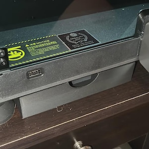 May include: A close-up of a black 3D printer with a pull-out drawer. The printer has a USB port and a small screen. There are warning labels and a logo on the top of the printer. The printer is sitting on a dark brown surface.