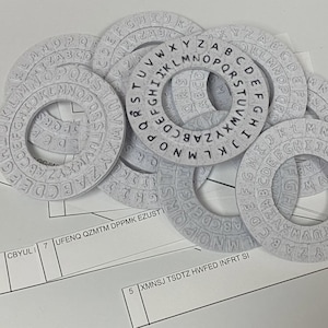 Op de afbeelding: Een verzameling lichtgrijze cijferwielen met gegraveerde letters en symbolen. De wielen zijn in een stapel gerangschikt, waarbij sommige elkaar overlappen. De afbeelding toont ook een vel papier met lijnen en tekst.