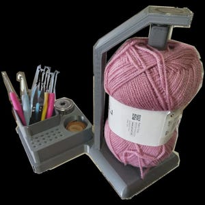 Può includere: Un organizer per filati grigio con una grande palla di filato rosa e vari uncinetti. L'organizer ha una sezione per gli attrezzi e un supporto per il filato. Il filato è avvolto con un'etichetta bianca.