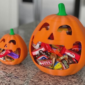Puede incluir: Dos calabazas naranjas impresas en 3D con caras talladas. La calabaza más grande está llena de caramelos envueltos, incluyendo Reese's, Twix y Kit Kat. La calabaza más pequeña también contiene caramelos.