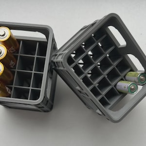 Puede incluir: Dos cajas de almacenamiento de baterías de plástico gris. Una caja contiene cuatro pilas AA doradas y plateadas. La otra caja contiene dos pilas AA. Las cajas tienen un diseño de rejilla y asas.