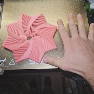 May include: A pink, star-shaped 3D-printed object with a spiral design. The object is on a gold-colored surface. The object's dimensions are approximately 6 inches in diameter.