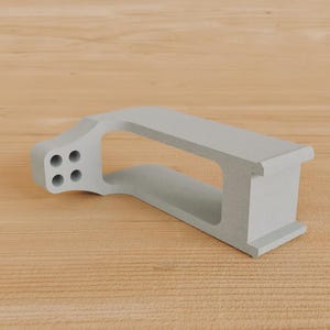 May include: A light grey, geometric object featuring four circular holes on one end. It has a rectangular aperture and rests on a wooden surface. The design is modern and minimalist.