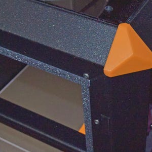 May include: Close-up of a black, textured corner of a structure with an orange, triangular accent piece. The structure has visible internal shelving and is secured with small screws. The orange piece adds a pop of colour.