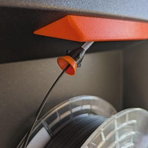 May include: An orange 3D-printed filament guide is mounted under a black surface. A black and gray filament is fed through the guide, leading to a spool of black filament. The guide is designed to help with 3D printing.
