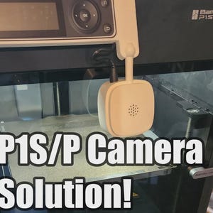 May include: A black 3D printer with a cream-coloured camera attachment. The camera is square with a speaker grill. The text "P1S/P Camera Solution!" is displayed in white block letters across the bottom of the image.