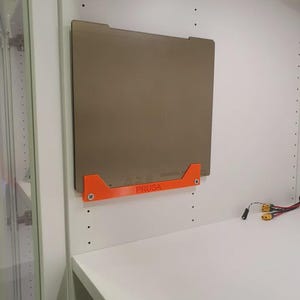 Könnte beinhalten: Eine rechteckige, beige 3D-Drucker-Bauplatte ist mit einer orangefarbenen Halterung an einer weißen Schrankwand befestigt. Auf der Halterung steht das Wort "PRUSA". Der Schrank hat ein weißes Regal.