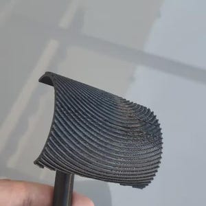 May include: A black, 3D-printed object with a handle and a curved, ridged top. The object is held in a person's hand. The ridges on the top are closely spaced and create a textured surface.