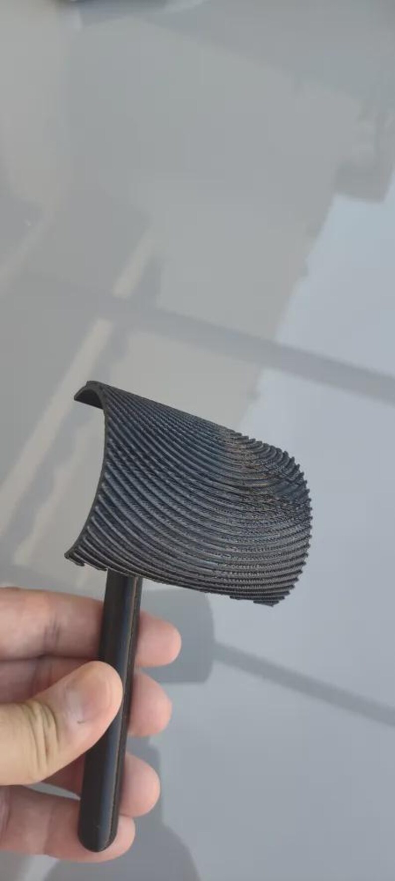 May include: A black, 3D-printed object with a handle and a curved, ridged top. The object is held in a person's hand. The ridges on the top are closely spaced and create a textured surface.