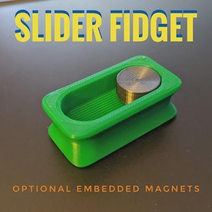 Puede incluir: Un juguete fidget deslizante verde con un disco plateado encima. El texto "SLIDER FIDGET" en amarillo y azul está encima del juguete, y "OPTIONAL EMBEDDED MAGNETS" en naranja debajo.