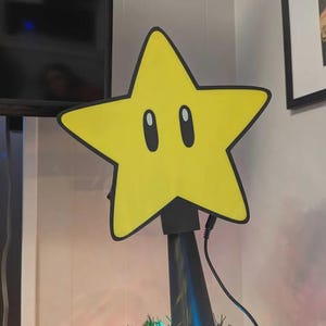 May include: A yellow star Christmas tree topper with black outlines and two oval eyes. The star is mounted on a black pole and sits atop a decorated Christmas tree with colourful lights.