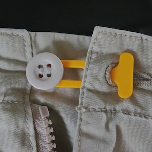 May include: Close-up of beige fabric with a white button and a yellow button extender. A yellow buttonhole tab is also visible. The image shows a zipper and stitching details, suggesting a garment.