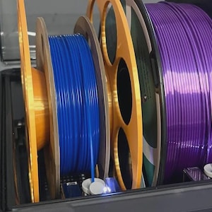 Peut inclure: Gros plan sur une unité de stockage de filaments pour imprimante 3D. L'unité contient plusieurs bobines de filaments de différentes couleurs, dont le bleu et le violet. Les bobines sont bien rangées et prêtes à l'emploi.