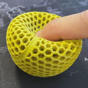 Könnte beinhalten: Ein hellgelbes, wabenförmiges 3D-gedrucktes Objekt. Ein Finger drückt in das Objekt. Das Design weist eine Reihe sechseckiger Zellen auf, die eine offene Struktur bilden. Die Farbe ist ein leuchtendes, sonniges Gelb.