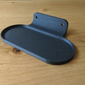 Puede incluir: Un estante negro para montar en la pared con una bandeja curva de forma ovalada y una placa trasera con dos orificios de montaje. El estante está hecho de un material texturizado y se encuentra sobre una superficie de madera.