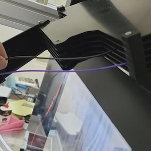 May include: A hand manipulating black and purple wires connected to a black metal structure. The background shows white tubes and other components, indicating an electronic or mechanical assembly.