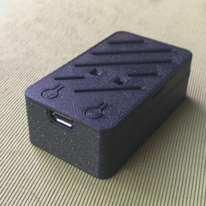 以下が含まれることがあります： テクスチャ加工された表面とUSBポートを備えた、小型の長方形の黒いデバイス。上部には斜めの溝と装飾的なシンボルがあります。このデバイスはおそらく、小型の電子部品またはハウジングです。