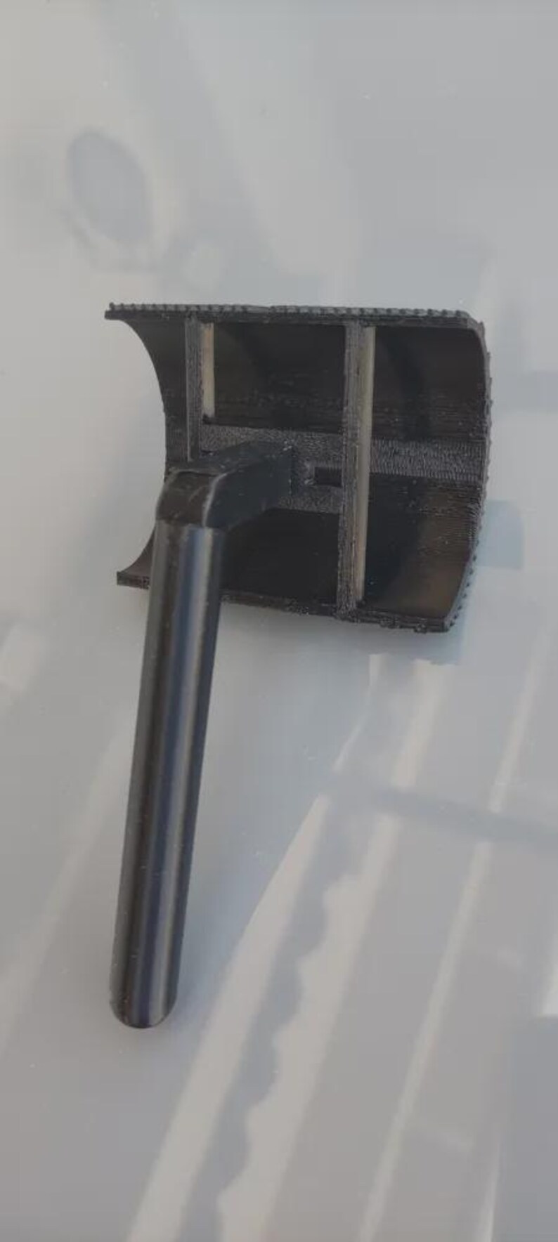 May include: A black 3D-printed object with a cylindrical handle and a rectangular head. The head has a curved top and internal supports. The handle is metallic gray.