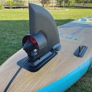 Può includere: Un paddleboard con un motore nero attaccato. La tavola ha un motivo a venature di legno chiaro con accenti blu e turchese. Il motore ha un interno rosso e un'elica nera. La tavola è su una superficie erbosa verde.