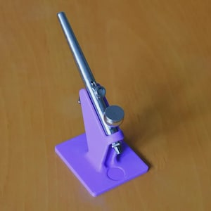 May include: A silver airbrush rests on a purple stand. The stand is rectangular with a triangular support and a circular indentation. The airbrush is metallic and angled, suggesting it's ready for use. The background is a warm, wooden surface.