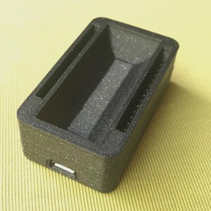May include: A rectangular, black 3D-printed object with a textured surface. It features a recessed interior with a ridged base and two parallel slots along the sides. A small rectangular opening is visible on one end.