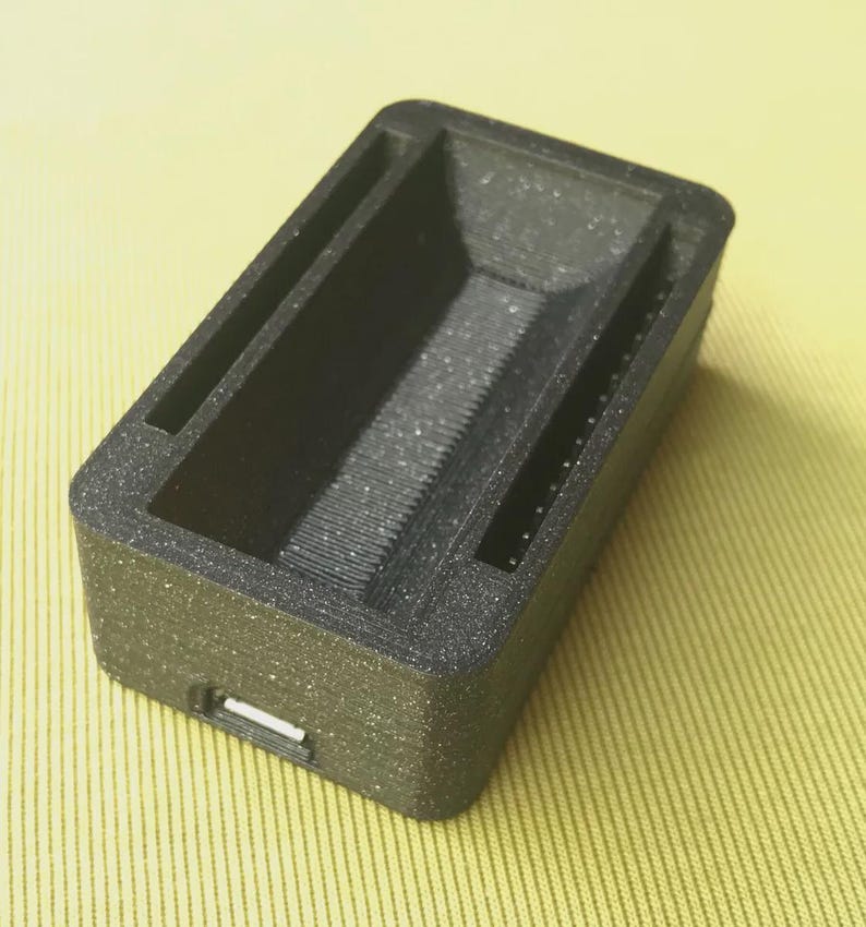 May include: A rectangular, black 3D-printed object with a textured surface. It features a recessed interior with a ridged base and two parallel slots along the sides. A small rectangular opening is visible on one end.