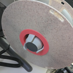 Könnte beinhalten: Nahaufnahme eines 3D-Drucker-Filament-Spulenhalters. Die Spule ist hellgrau gesprenkelt mit einem rosa Ring und einem weiß-grauen Mechanismus. Der Halter ist an einem schwarz-grauen Rahmen befestigt.