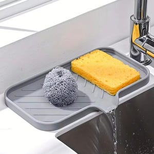 May include: A gray silicone sink caddy holds a yellow sponge and a gray metal scrubber. The caddy is positioned near a stainless steel faucet, with water flowing into the sink. This kitchen accessory helps organize cleaning supplies.