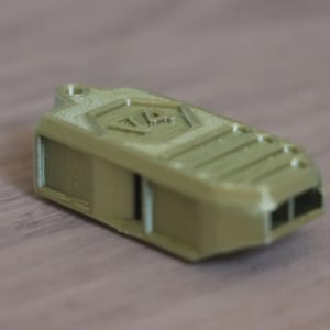 May include: A gold-coloured whistle with a rectangular body and a small loop for a lanyard. The whistle has a textured surface and a design on the top. It features two rectangular openings on one end.