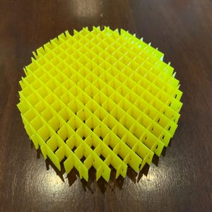 Puede incluir: Un objeto circular amarillo brillante, similar a una rejilla. El objeto está hecho de piezas plegadas interconectadas, creando una estructura tridimensional y abierta. El diseño es simétrico y geométrico, con un patrón repetitivo de aberturas cuadradas. El objeto está sobre una superficie marrón oscura.