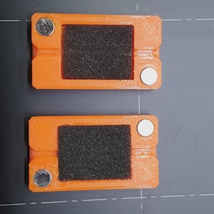 Puede incluir: Dos objetos rectangulares naranjas con un centro texturizado negro y un imán circular plateado y blanco en cada extremo. Los objetos están colocados sobre una superficie gris.