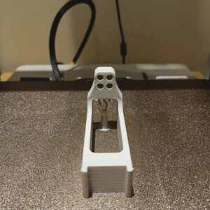 May include: A white 3D-printed component with a rectangular base and a detailed top section with four circular holes. The object has a central support structure, possibly for a larger assembly.