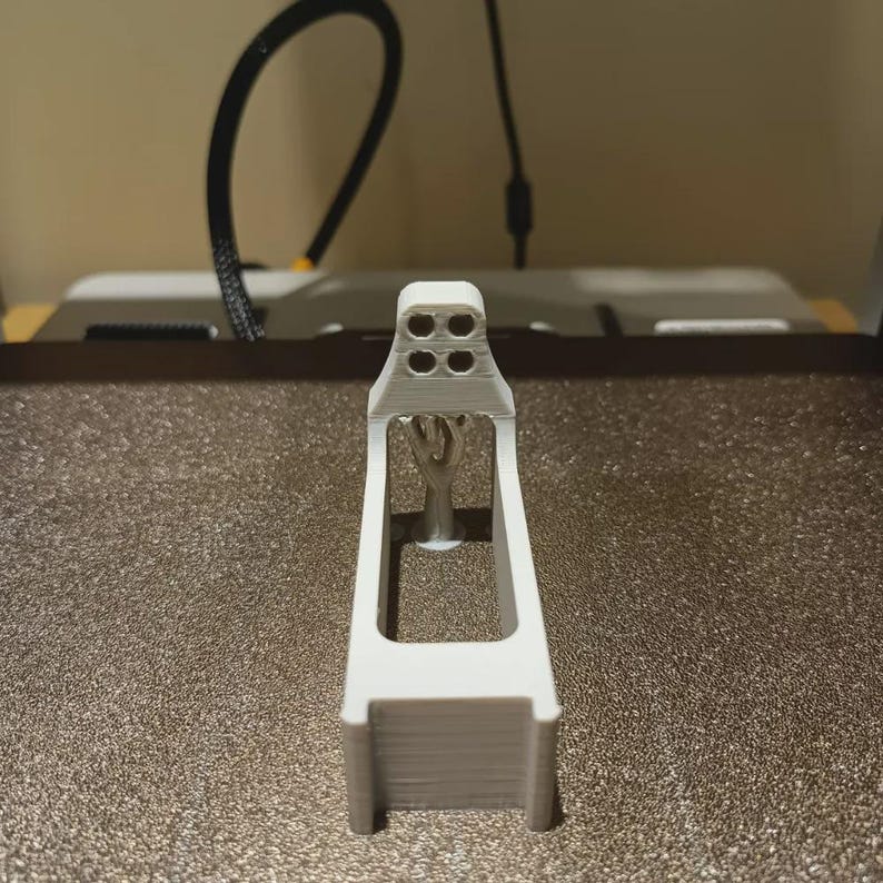 May include: A white 3D-printed component with a rectangular base and a detailed top section with four circular holes. The object has a central support structure, possibly for a larger assembly.