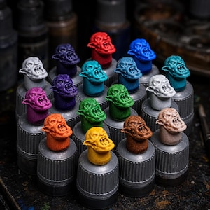 May include: A collection of miniature orc heads in various solid colours including red, blue, teal, purple, green, orange, yellow, brown, white, and beige. Each head is mounted on a grey cylindrical base, displaying sharp teeth and pointed ears.