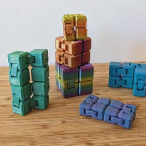 May include: Several colourful fidget cubes in shades of green, blue, purple, and orange. The cubes are arranged in different ways, some stacked and others laid out flat. The background is a light wood surface.