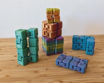 May include: A collection of colorful fidget cubes in shades of green, blue, purple, and orange. The cubes are arranged in various configurations, some stacked and others laid out flat. The background is a light wood surface.